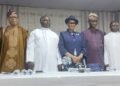 Eight dignitaries in formal and traditional attire pose for a group photo at a ceremony, behind a table with water bottles.