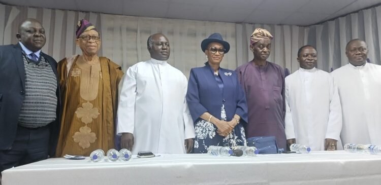 Bishop-Joseph-Akinfenwa_copy_800x365_1 - Newscoven Eight dignitaries in formal and traditional attire pose for a group photo at a ceremony, behind a table with water bottles.