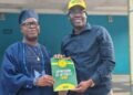 Two men pose holding an 'Expression of Interest Form 2026' booklet; one wears traditional blue attire and cap, the other a blue shirt and yellow cap.