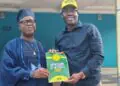 Two men pose holding an 'Expression of Interest Form 2026' booklet; one wears traditional blue attire and cap, the other a blue shirt and yellow cap.
