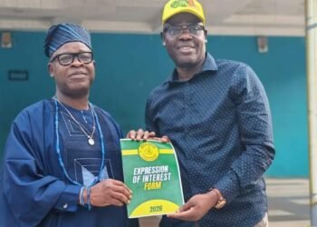 Two men pose holding an 'Expression of Interest Form 2026' booklet; one wears traditional blue attire and cap, the other a blue shirt and yellow cap.