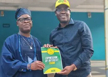 Two men pose holding an 'Expression of Interest Form 2026' booklet; one wears traditional blue attire and cap, the other a blue shirt and yellow cap.