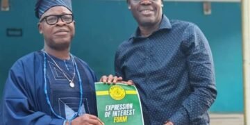 Two men pose holding an 'Expression of Interest Form 2026' booklet; one wears traditional blue attire and cap, the other a blue shirt and yellow cap.