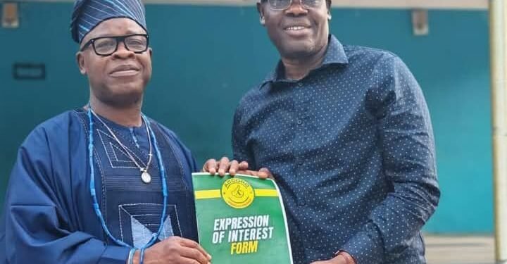 Two men pose holding an 'Expression of Interest Form 2026' booklet; one wears traditional blue attire and cap, the other a blue shirt and yellow cap.