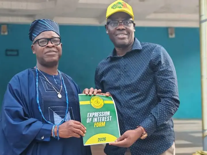 Two men pose holding an 'Expression of Interest Form 2026' booklet; one wears traditional blue attire and cap, the other a blue shirt and yellow cap.